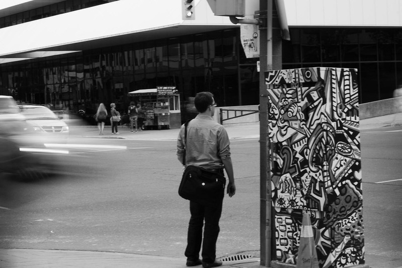 A person standing at an intersection. The person has its back to the camera, and the cars are passing by, in a motion blur effect.